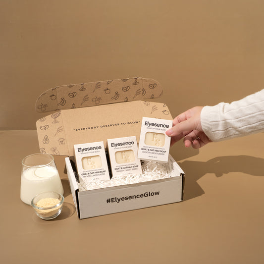 Hand placing an Elyesence Goat & Oat Milk Soap bar into a gift box with three soaps, glass of milk and oats beside it, with hashtag #ElyesenceGlow on the box