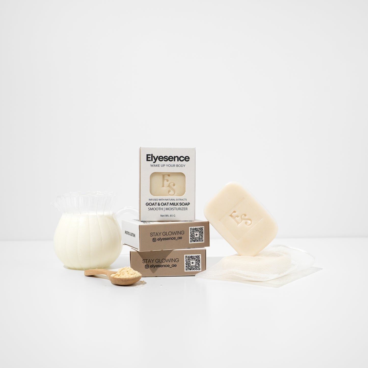 Goat & Oat Milk Soap by Elyesence displayed with product packaging, mesh soap bag, a jug of milk, and a wooden spoon of oat powder on a clean white background