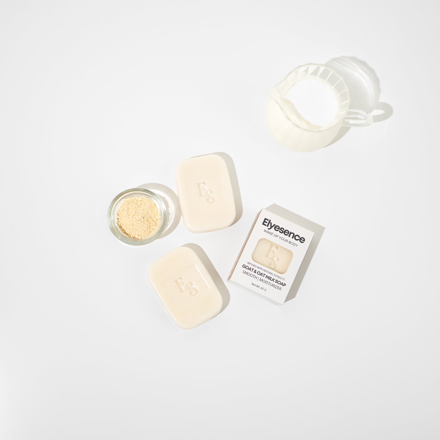 op-down view of Elyesence Goat & Oat Milk Soap with two unboxed soap bars, one boxed bar, a glass bowl of oat powder, and a jug of milk on a white background