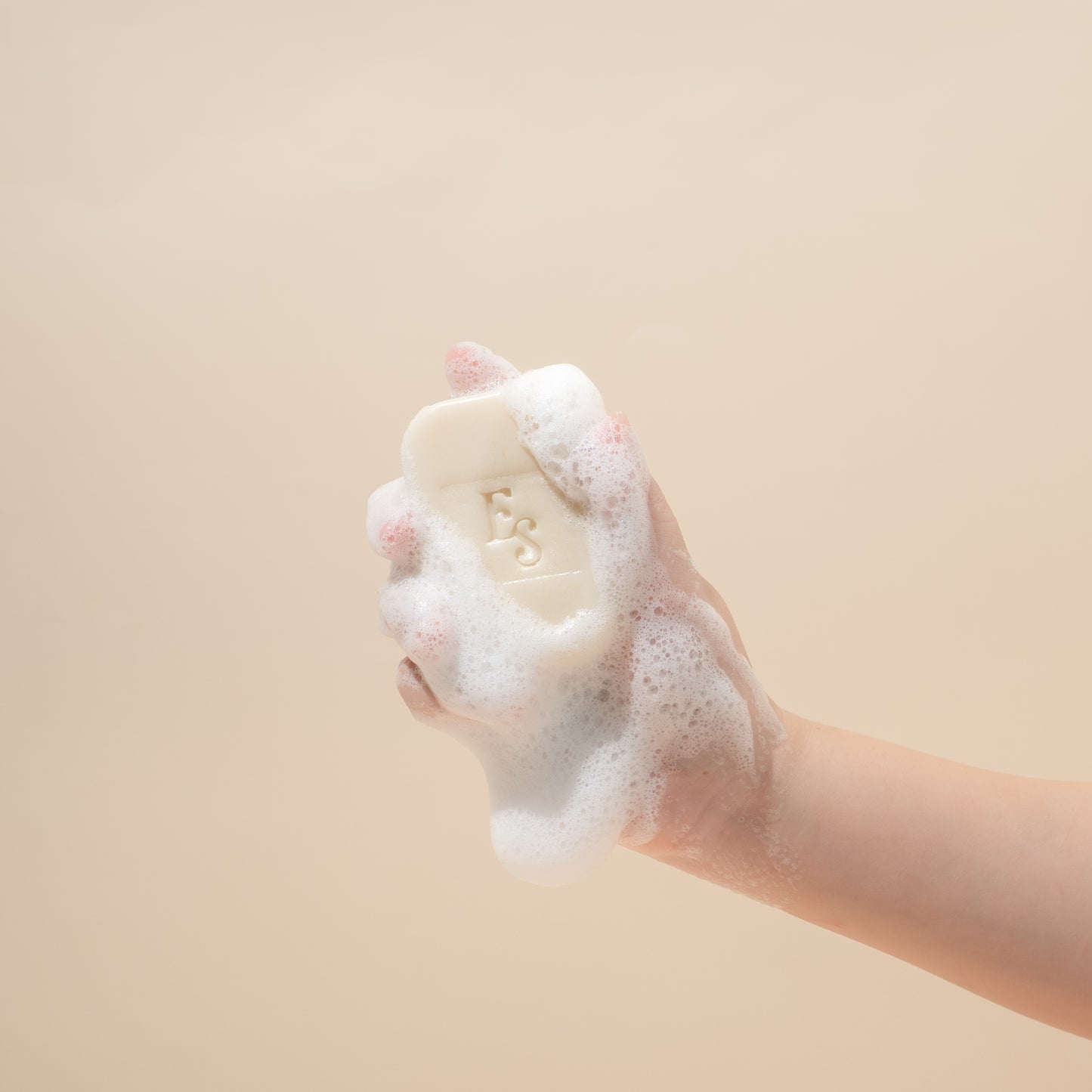 Hand holding a lathered Elyesence Goat & Oat Milk Soap bar with rich foam against a soft beige background
