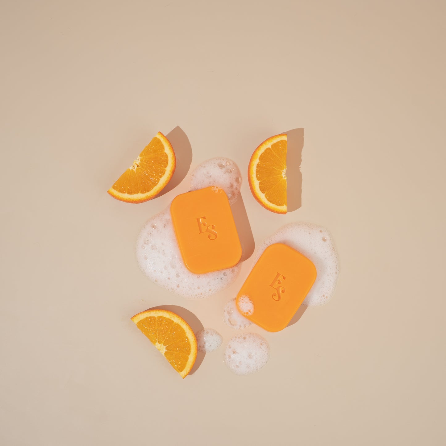 Two Elyesence Orange Radiance Soaps with bubbles and orange slices in a clean flat lay on a beige background