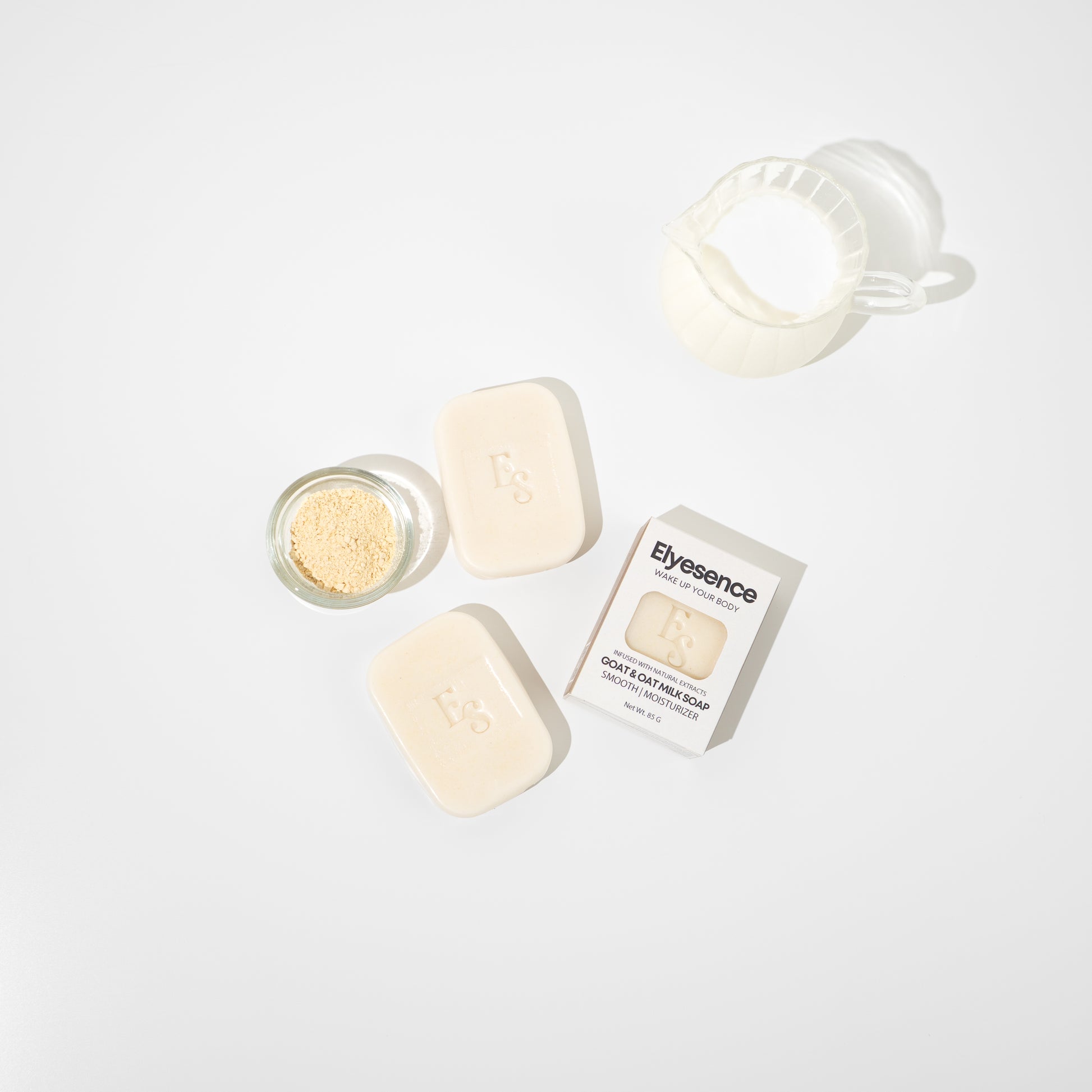 op-down view of Elyesence Goat & Oat Milk Soap with two unboxed soap bars, one boxed bar, a glass bowl of oat powder, and a jug of milk on a white background